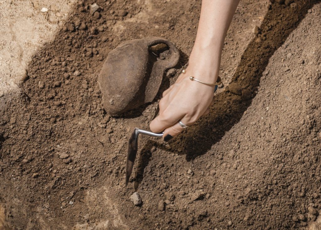Archaeologist digging with hand trowel, recovering ancient pottery object from an archaeological site.